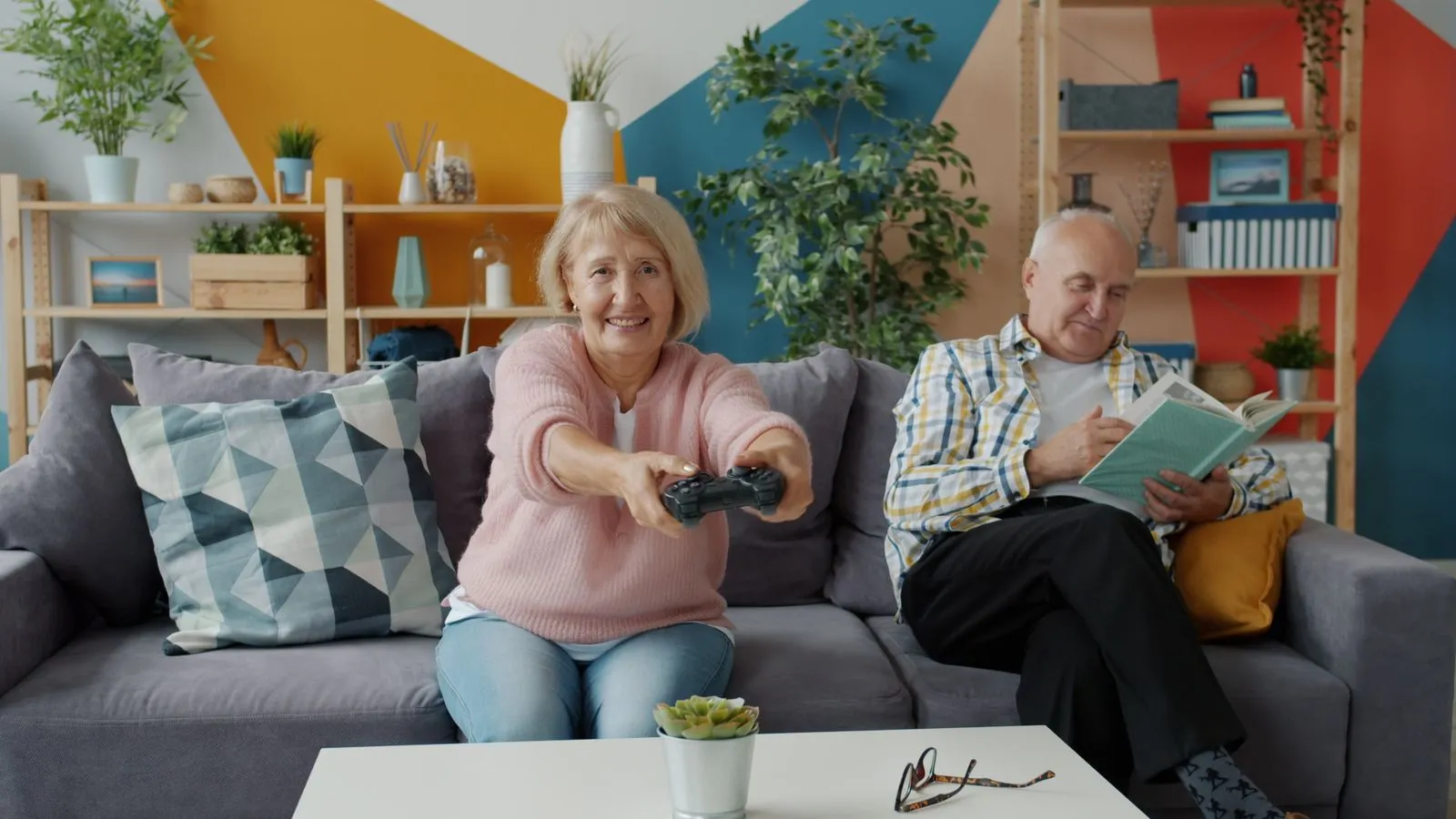 Elderly couple enjoying hobbies—gaming and reading—on a cozy home couch.